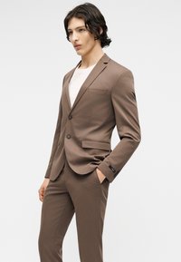 Άνδρας με σκούρα μαλλιά φορώντας εφαρμοστό καφέ κοστούμι και λευκό πουκάμισο, στέκεται με το ένα χέρι στην τσέπη μπροστά σε ένα μονόχρωμο φόντο.