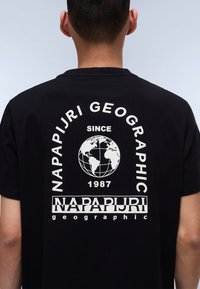 Juodas marškinėliai su balta užrašu "NAPAPIJRI GEOGRAPHIC" ir žemėlapiu dizaino nugaroje, po žemėlapiu esantis metai "1987".