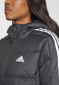 Giacca imbottita nera con cappuccio, caratterizzata da strisce bianche sulle spalle. Include una chiusura frontale con zip e un logo Adidas sul petto.