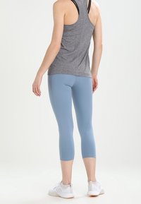 Světle modré capri legíny vyrobené z elastického materiálu, v kombinaci se šedým tílkem a bílými sportovními botami, viděné zezadu.