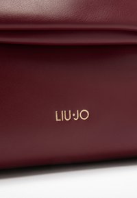 Close-up van een bordeauxleren tas met het gouden LIU·JO-logo dat op het oppervlak is geperst, waarbij de textuur en een gevouwen ontwerpelement worden getoond.