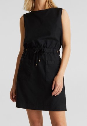 Robe noire sans manches avec taille à cordon, boutons sur les côtés et deux poches à l'avant, portée par une personne aux cheveux blonds.