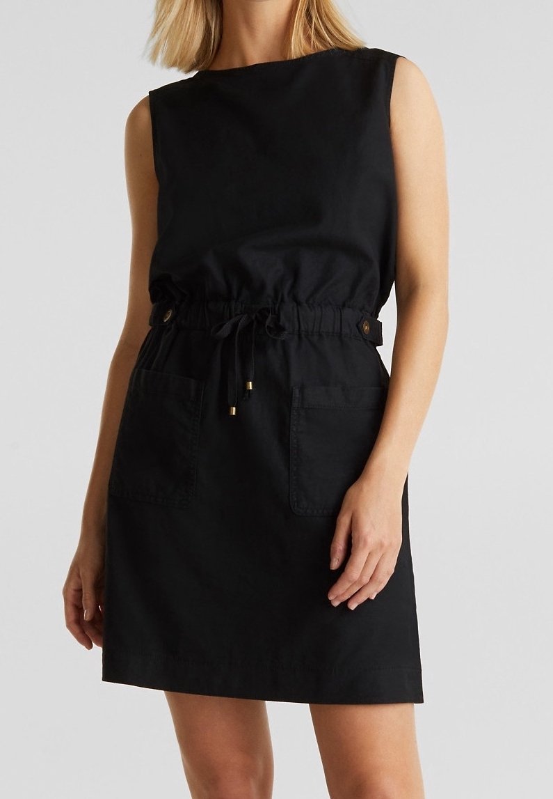 Robe noire sans manches avec taille à cordon, boutons sur les côtés et deux poches à l'avant, portée par une personne aux cheveux blonds.