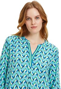 Lichtblauwe blouse met een geometrisch patroon in groen, blauw en wit. Heeft een V-hals met een contrasterende gestreepte afwerking en lange mouwen.