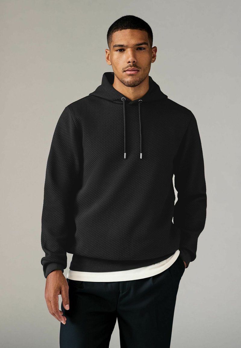 Zwarte hoodie van textuurstof, voorzien van een trekkoordcapuchon en geribbelde mouwen. Draagt over een lichtgekleurde shirt met een donkere broek.
