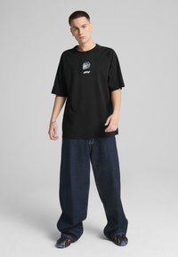 Black oversized t-shirt with a round neckline and logo graphic on the chest, paired with loose-fitting dark denim pants.