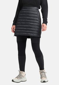 Zwarte puffer rok met horizontale stiksels, semi-doorzichtig materiaal en ritsen aan de zijkanten, gecombineerd met zwarte leggings en lichte wandelschoenen.