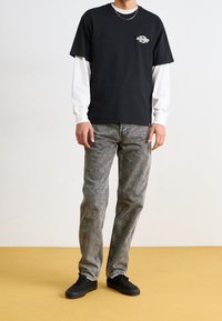 Černé tričko s krátkým rukávem přes bílé tričko s dlouhým rukávem, doplněné o šedé džíny a černé tenisky. Na hrudi je logo.