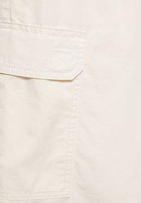 Close-up of beige fabric showing a stitched rectangular pocket with a flap and reinforced stitching detail on the corner.
