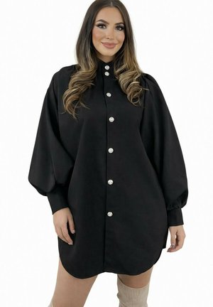 Femme portant une robe noire à manches longues boutonnée avec des manches bouffantes, souriante, aux longs cheveux ondulés et chaussettes beige montant jusqu'au genou.