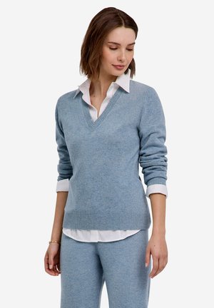 Femme regardant vers le bas, portant un pull bleu clair à col en V sur une chemise blanche avec col et un pantalon bleu clair assorti sur un fond blanc uni.