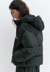 Mladá žena s tmavými vlnitými vlasmi nosí veľkú čiernu prešívanú bundu s kapucňou, ruky v náprsných vreckách, dívajúca sa dole na jednoduchom pozadí.