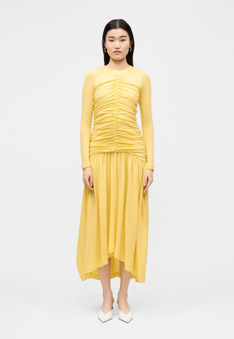 Femme aux longs cheveux foncés debout portant un top jaune à manches longues froncé et une jupe midi fluide assortie avec des talons blancs pointus.