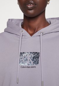 Jasnofioletowa bluza z kapturem z naszywką z logo z przodu, zawierającą metaliczne akcenty. Zamykanie na sznurki z metalowymi końcówkami i miękka tekstura materiału.