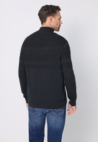 Suéter de punto gris oscuro con cuello alto y rayas texturizadas, combinado con jeans de mezclilla azul, visto desde atrás.