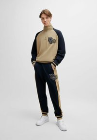 Completo sportivo a due pezzi con una giacca beigem con zip e maniche blu navy, abbinato a pantaloni blu navy. Entrambi presentano loghi ricamati e strisce.