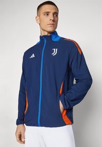 Giacca sportiva blu navy con accenti arancioni sui lati, zip blu e logo bianco. Realizzata in materiale leggero, presenta un colletto alto.