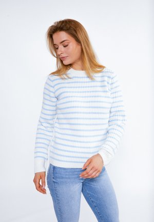 Woman with light brown hair wearing white sweater with light blue horizontal stripes and light blue jeans, looking down with relaxed expression.