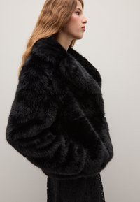 Giacca in pelliccia sintetica nera con colletto alto, morbido al tatto e maniche lunghe, abbinata a un indumento in pizzo sottostante. Design semplice.