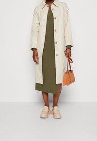 Persona vestida con un abrigo beige sobre un vestido verde oliva con una abertura lateral, sujetando un pequeño bolso de ante marrón claro y unos mocasines beige de tacón grueso.