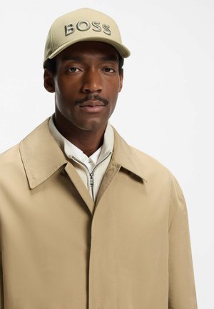 Beige jacket with a broad collar over a light gray zip-up sweater. Cream baseball cap with "BOSS" embroidered in bold letters.