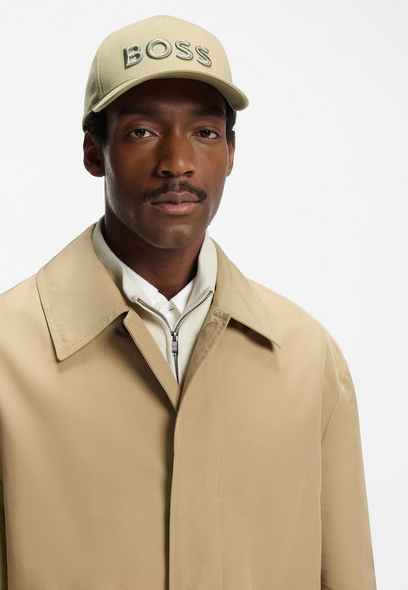 Beige jacket with a broad collar over a light gray zip-up sweater. Cream baseball cap with "BOSS" embroidered in bold letters.