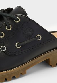 Chaussure en cuir noir avec œillets pour lacets, semelle épaisse en caoutchouc marron avec des rainures profondes et logo arbre embossé sur le panneau latéral.