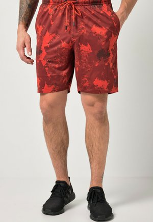 Mand iført røde og sorte mønstrede træningsshorts med snøre, sorte sneakers og hænder ved siden af kroppen mod ensfarvet baggrund.