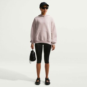 Ung kvinde iført en pink hættetrøje, sorte cykelshorts, sort kasket, solbriller og med en lille sort håndtaske, stående på en hvid baggrund.