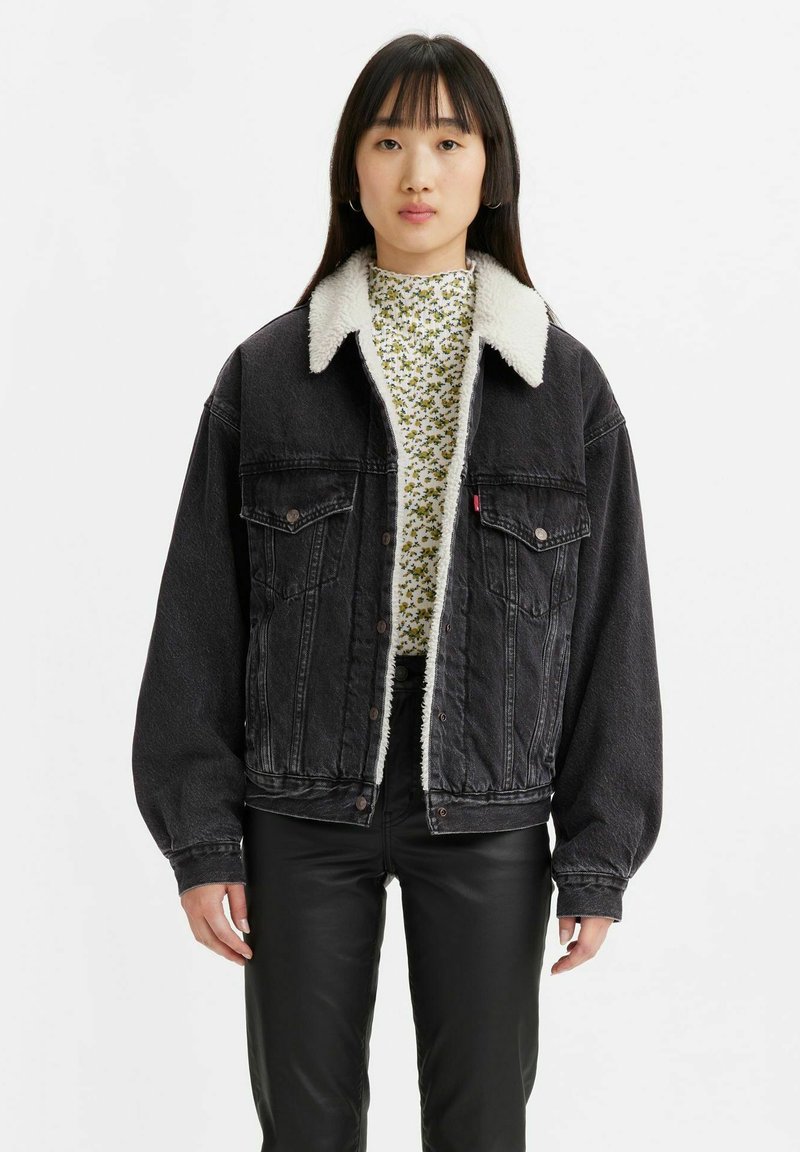 Giacca in denim nero con foderatura in sherpa, vestibilità oversize, due tasche frontali e chiusura con bottoni. Indossata sopra un top bianco a motivi floreali.