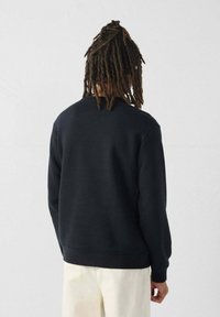 Sudadera negra con cuello redondo, puños y dobladillo acanalados. Presenta una textura suave y un corte relajado, llevada sobre pantalones de color claro.