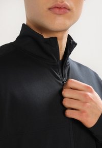 Chaqueta negra con cremallera, hecha de un tejido texturizado. Presenta un cuello alzado y una cremallera en el cuello. La imagen de cerca muestra una mano agarrando la cremallera.