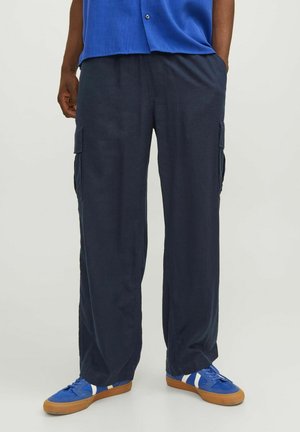 Unterkörper mit lockeren dunkelblauen Cargohosen mit Seitentaschen, blauen Sneakers mit weißen Streifen und einem blauen Hemd, das in der Taille gesteckt ist.