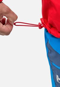 Serrer à la main le cordon rouge avec un stoppeur en plastique sur une veste rouge portée sur un pantalon bleu et marine.