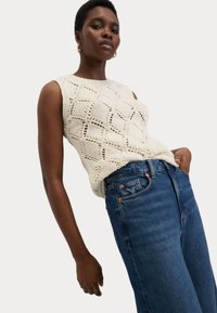 Gebreid mouwloos topje in crème met een diamantpatroon, gecombineerd met blauwe high-waisted jeans met zichtbare stiksels en zakken.