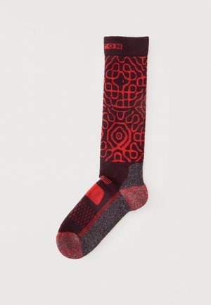 Calcetín hasta la rodilla con patrones geométricos rojos sobre fondo oscuro, con talón y puntera reforzados en rojo y gris.