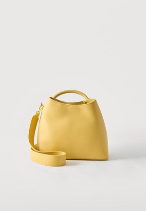Sac à main en cuir texturé jaune avec une courte poignée et une longue bandoulière amovible, debout sur fond blanc.