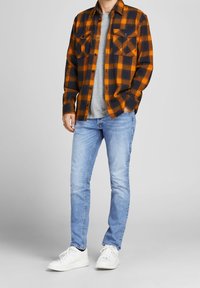 Man som bär en orange och svart rutiga skjorta över en grå t-shirt, ljusblå jeans och vita sneakers, står med ena handen i fickan.