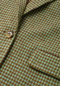 Veste à motif pied-de-poule marron, vert et bleu avec un bouton et une poche à rabat visibles sur un tissu texturé.