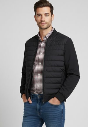 Chaqueta bomber negra acolchada con mangas largas, puños acanalados y un tejido suave. Lleva puesta sobre una camisa de cuadros y combinada con jeans azules.