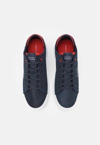 Marineblaue, strukturierte Sneaker mit roten Akzenten, einer weißen Sohle und Schnürsenkeln sowie einem Etikett mit "Tommy Hilfiger" auf der Zunge.