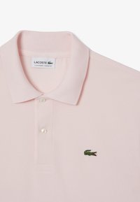 Ljusrosa polo-shirt gjord av texturerat tyg, med tre knappar vid kragen och en liten grön krokodil-logga på vänster bröst.