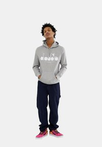 Felpa grigia con logo bianco, pantaloni larghi blu scuro e sneakers rosa con lacci neri. Il modello ha i capelli ricci e le mani in tasca.