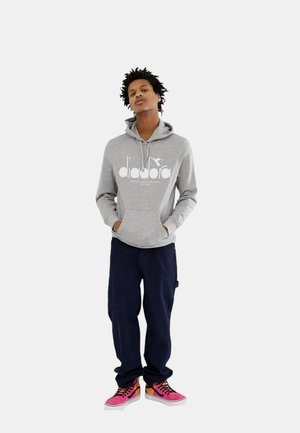 Felpa grigia con logo bianco, pantaloni larghi blu scuro e sneakers rosa con lacci neri. Il modello ha i capelli ricci e le mani in tasca.