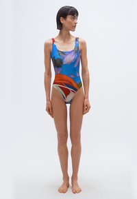 Γυναίκα με κοντά σκούρα μαλλιά, φορώντας πολύχρωμο ολόσωμο μαγιό με λουλούδια, όρθια ξυπόλυτη μπροστά σε απλό λευκό φόντο, κοιτάζοντας πλάγια.