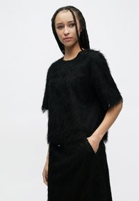 Czarna bluzka z sztucznego futra z krótkimi rękawami i okrągłym dekoltem, zestawiona z dopasowaną czarną spódnicą. Tkanina ma miękką, kudłatą teksturę.