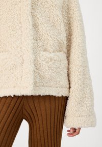 Beige fluffig fleece-pullover med texturerad yta, med framfickor och vida ärmar, som är ihop med ribbade bruna stickade leggings.