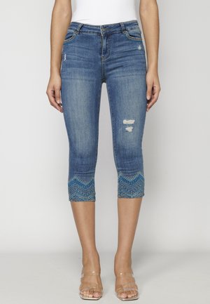 Mujer que lleva vaqueros cortos de mezclilla azul desgastados con dobladillos estampados y sandalias con tiras transparentes, de pie sobre un fondo liso.