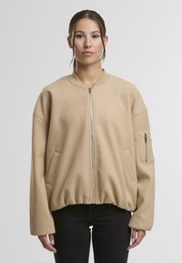 Béžová bomber bunda vyrobená z fleecu s předním zipem, žebrovanými manžetami a dvěma bočními kapsami. Obsahuje kapsu na zip na rukávu pro uložení.