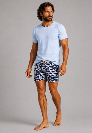 Mann mit dunklem lockigem Haar und Bart, der barfuß geht und ein hellblaues T-Shirt sowie marineblaue und weiße Shorts mit Rautenmuster trägt.